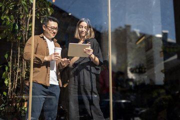 Multiracial business people watching something on digital tablet inside unknown building. Concept of remote and freelance work. Idea of teamwork. Caucasian girl in earphones. Asian man with coffee cup