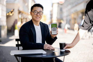 Asian man show smartphone with app of Coronavirus Vaccination to female waiter at outdoor cafe. Concept of health protection during pandemic. Idea of rest and leisure. Girl put coffee on client table