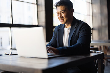 Asian businessman typing on laptop during work in cafe. Concept of remote and freelance work. Smiling adult successful man wearing suit and glasses sitting at wooden desk. Sunny day