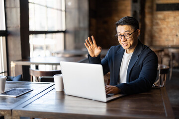 Asian businessman having a video call on laptop in cafe. Concept of remote and freelance work. Smiling adult successful man wearing suit and glasses sitting at wooden desk. Sunny day