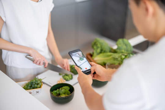 Man taking video on smartphone of woman cooking salad from fresh vegetables and fruits. Concept of video blogging. Idea of healthy and vegetarian eating. Partial image of couple at home kitchen