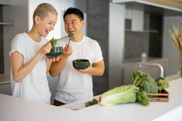 Laughing multiracial couple eating salad on kitchen at home. Concept of healthy and diet eating. Idea of domestic lifestyle. Asian man and caucasian woman at table with organic vegetables and fruits