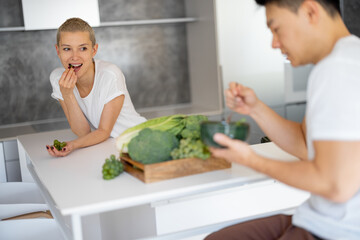 Asian man holding bowl with salad while his girlfriend eating grapes and looking on him. Concept of healthy and vegetarian eating. Domestic lifestyle. Multiracial couple on kitchen in studio apartment