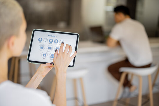 Woman pushing button on kitchen app of smart home on digital tablet at home. Concept of modern home control. Idea of domestic lifestyle. Cropped image of girl. Blurred man on background using laptop