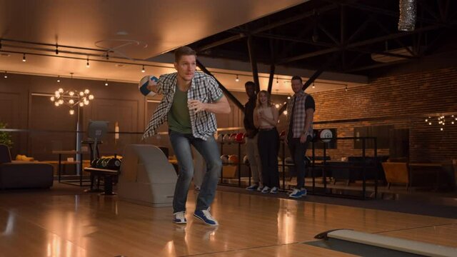 A man in a bowling alley throws a ball on the track and knocks out a shot in slow motion and jumps and dances for joy. A group of multi-ethnic friends play bowling together clapping happy, supporting