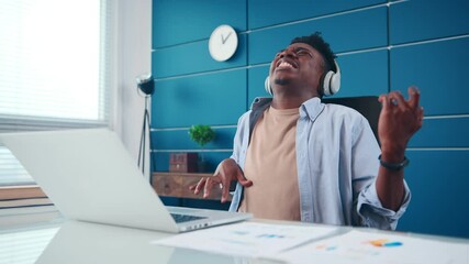 Cheerful African American man in wireless headphones pretends to play guitar. Passionate Hispanic male listens to music sits at office desk, dancing singing along. Young freelancer lifestyle concept.