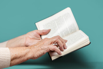 Wrinkled hands of a senior woman are holding a book. Turquoise background. The concept of education and reading