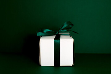 A small gift with a green ribbon and green christmass ball on green background. White gifts with a green bow. Christmas gifts. Gift for woman. Holiday background with gift, white satin 