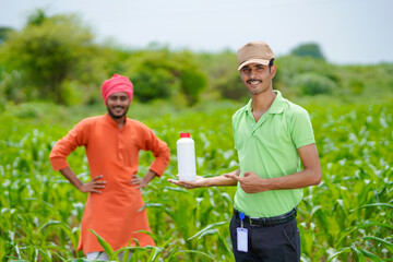 Young indian agronomist holding liquid fertilizer bottle with farmer at green agriculture field.