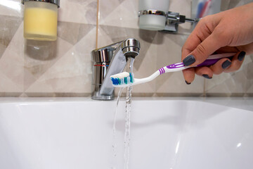 A woman's hand holding a toothbrush and toothpaste under running water from the tap.