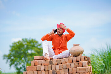 Indian farmer sitting on bricks and talking on smartphone at green corn field.