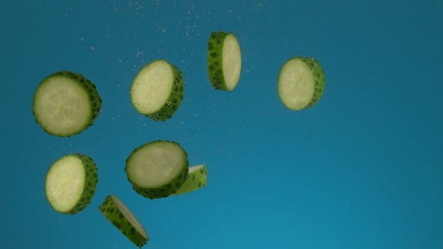 Slow motion fresh juicy green sliced cucumber falls into the water with splashes and bubbles. Vegetable on a blue background.