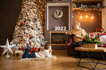 Grandmother reading book to grandchildren near Christmas tree at home in New Year 2022
