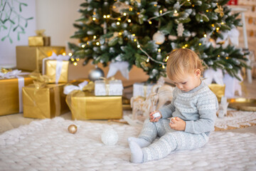 Obraz na płótnie Canvas Adorable baby girl sitting near Christmas tree with festive lights and xmas gifts. Chrismas and New Year.