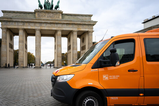 Two rickshaw bike taxis stand in front of the An orange BSR garbage truck in front of the Brandenburg Gate