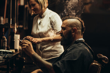 Young bearded man with a mohawk getting haircut by hairdresser and smoking a cigarette