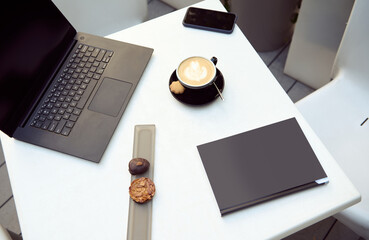 Flat lay composition of delicious pastries on a white table next to a laptop and a cup of delicious aromatic cappuccino coffee, a mobile phone and a black hardcover book. Copy ad space