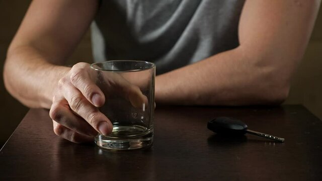 Tipsy man wearing grey t-shirt drinks whiskey from glass to bottom and takes car key deciding to drive auto after alcohol extreme closeup.
