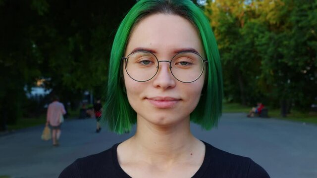 Portrait of nice girl with green short hair in glasses. Young woman looking at camera, smiling, blinking in city park. Gaze of lady in black with grey blue eyes. Front view