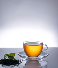 Fragrant hot tea in a transparent cup on a saucer. Mint leaves and tea leaves in the foreground. Light mirrored table