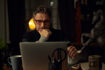 Older man sitting at desk in dark cosy room working on laptop computer in home office. Mature age, middle age, mid adult casual man in 50s, confident happy smiling.