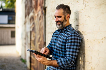 Portrait of modern businessman with beard using digital tablet while standing in front of brick wall outdoor.	