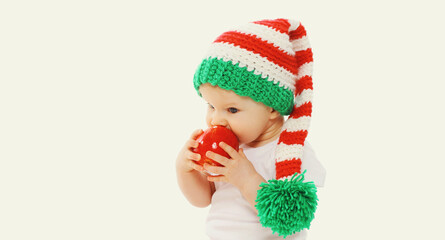 Portrait of little baby in knitted gnome winter hat biting red apple on a white background