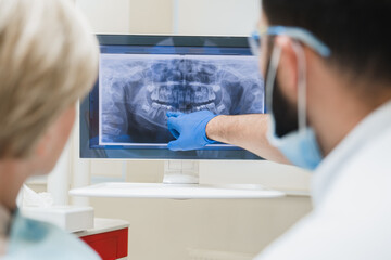 Back view shot of male dentist orthodontist showing explaining an x-ray photo of jaws on computer screen to a female patient in dental clinic. Stomatology and healthcare concept