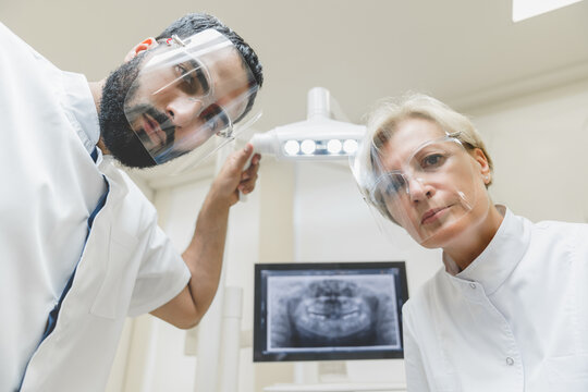 Two stomatologists assistant orthodontists dentists looking checking up patient`s mouth cavity, curing teeth tooth in protective masks. Low angle view