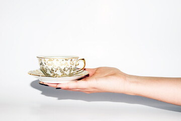 Mano sosteniendo una pequeña taza de té fina con adornos de flores doradas pintadas a mano sobre...