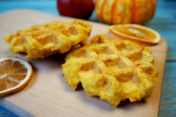a close-up of Belgian gluten-free and lactose-free pumpkin waffles lie on a wooden board next to slices of dried orange and a small pumpkin on a blue wooden table background. side view