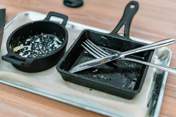 Empty black frying pans stand inside a stainless steel tray on a table. Restaurant. Dirty. Mess. Finished. Food. Restaurant. Cafe. Fork and knife