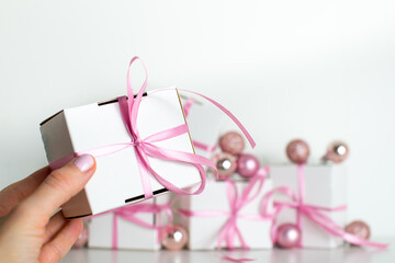 A female hand holds a small gift with a pink ribbon on white background. White gifts with a pink bow. Christmas gifts. Gift for woman. Holiday pink background with gift, white satin bow, ribbon. 