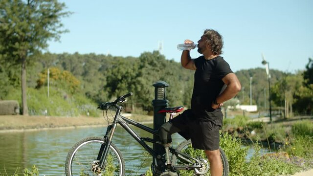 Long shot of man with bionic leg drinking water after cycling. Side view of tired person standing in park next to river with artificial leg on bicycle and having break. Disability, recovery concept
