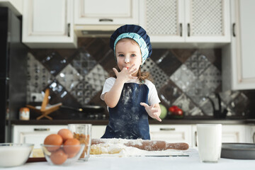 A little girl in an apron and a chef's hat sprinkle the dough with flour in the kitchen.