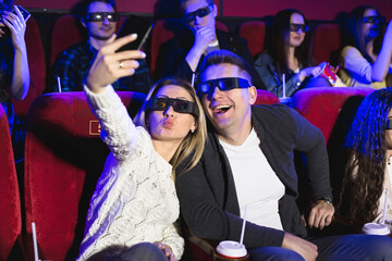 Beautiful girl and guy laughing happily making a selfie together during a movie at the cinema. Young couple watching a movie at a cinema and photographing themselves with a smartphone.