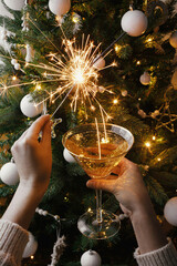 Happy New Year! Hands holding burning sparkler and champagne glass against christmas tree lights
