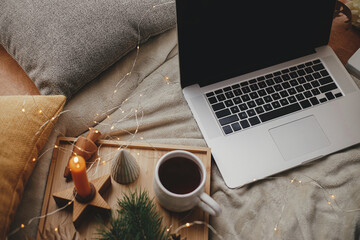 Laptop on soft bed, cup of tea, christmas lights, stars, pine, candle, pillows. Freelance and blog