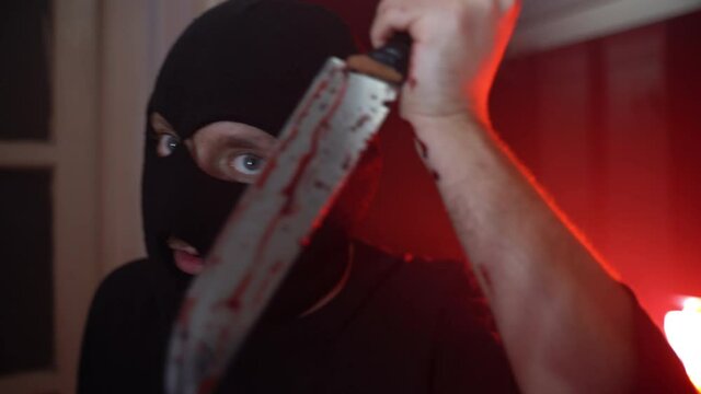 Portrait of spooky serial killer in black balaclava with demon eyes looking on camera and holding bloody knife in hands, psycho maniac ready for attacking victim with aggression and cold weapon. Cruel