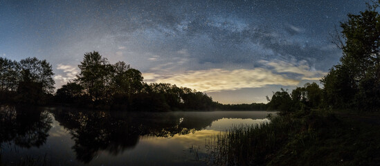 Panorama of Milky Way at night over a small lake with silhouette of trees on the shore line