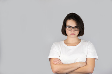 Fototapeta premium Studio photo on the background of a beautiful serious girl with dark hair in glasses with crossed hands on a white t-shirt