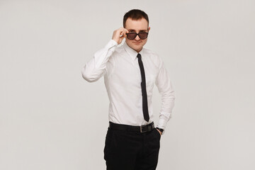 A young successful confident guy in a white shirt and a black tie adjusts his sunglasses on a gray background. Isolated with copy space