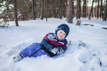 Fototapeta na wymiar Happy boy enjoys winter season and lies in the snow in forest on sunny winter day. Lovely winter scenery, active weekend, seasonal outdoor activities, happy family lifestyle