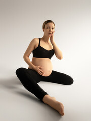 Fototapeta premium Pregnant smiling woman sitting on white floor in studio room. Looking at camera. Maternity. Motherhood. Young happy pregnant woman sitting. Surprised Pregnant woman. People concept