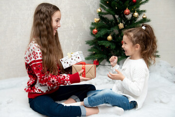 Two cute smiling girls sit next to the Christmas tree and give each other gifts. New Year. Christmas.
