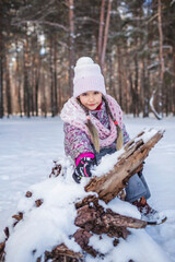 Happy girl in knitted hat enjoys winter season and sits in the snow in forest on sunny winter day. Lovely winter scenery, active weekend, seasonal outdoor activities, happy family lifestyle