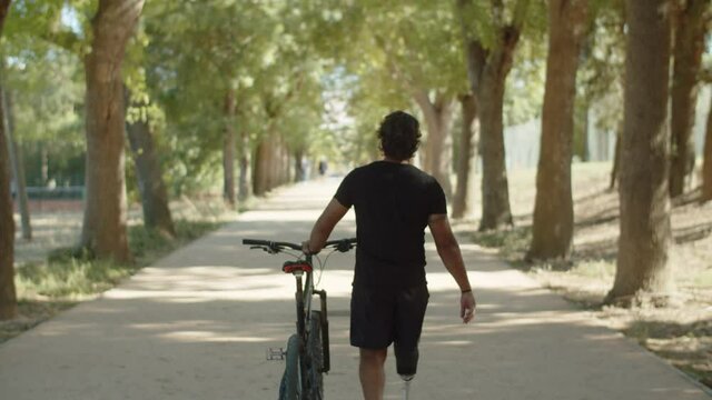 Back view of man with bionic leg walking down alley with bike. Long shot of athlete with artificial limb in sportswear spending time outdoor, having cardio training in park. Disability, sport concept