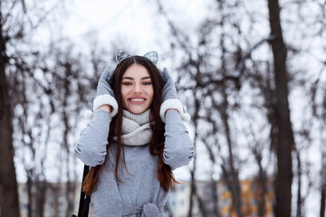 Pretty fashionable woman dressed in gray dress, gloves and cat headband outdoors in winter park. 