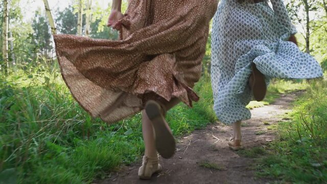 Girls in light dresses are running in a sunny forest. Girls' legs in close-up. The girls hold hands and run down the path from the camera. A shot from a steadicam. Mouse angle