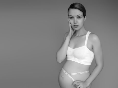 Beautiful pregnant young woman in clothes for pregnant women is measuring her bare tummy, smiling, on a background. Picture of happy pregnant woman posing over wall. Looking at camera